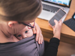 Mom looks down at baby in sling, with a phone in her hand and laptop on desk 