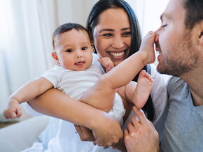 Mom holds baby, smiling. Baby has foot on dad’s nose.