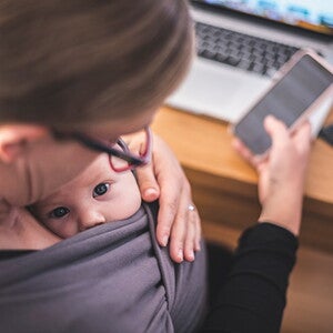 Mom looks down at baby in sling, with a phone in her hand and laptop on desk Mom looks down at baby in sling, with a phone in her hand and laptop on desk