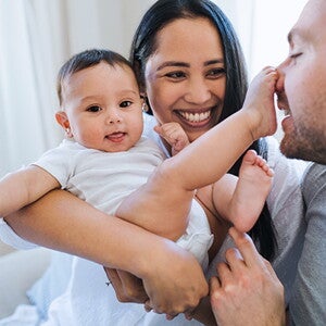 Mom holds baby, smiling. Baby has foot on dad’s nose. Mom holds baby, smiling. Baby has foot on dad’s nose.